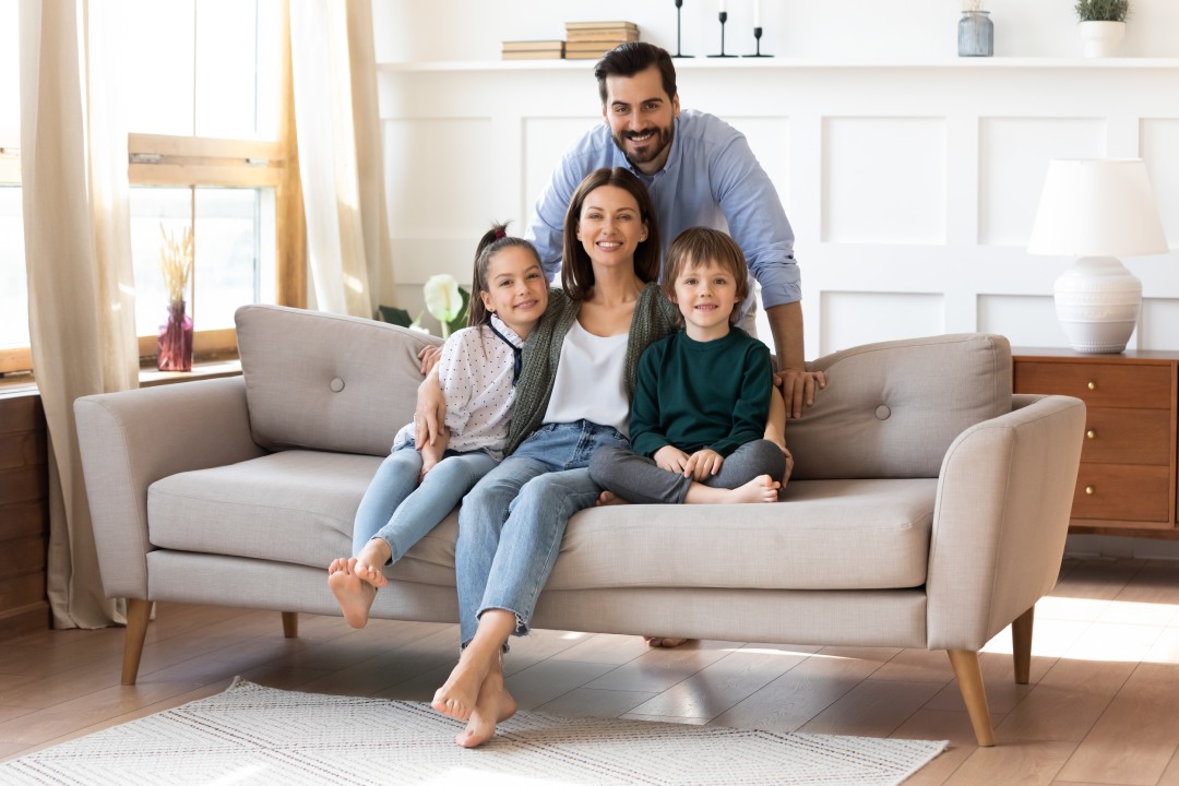 Portrait of smiling bearded father standing near couch with sitting happy wife and little children. Joyful affectionate family of four posing for photo, looking at camera, good relations concept